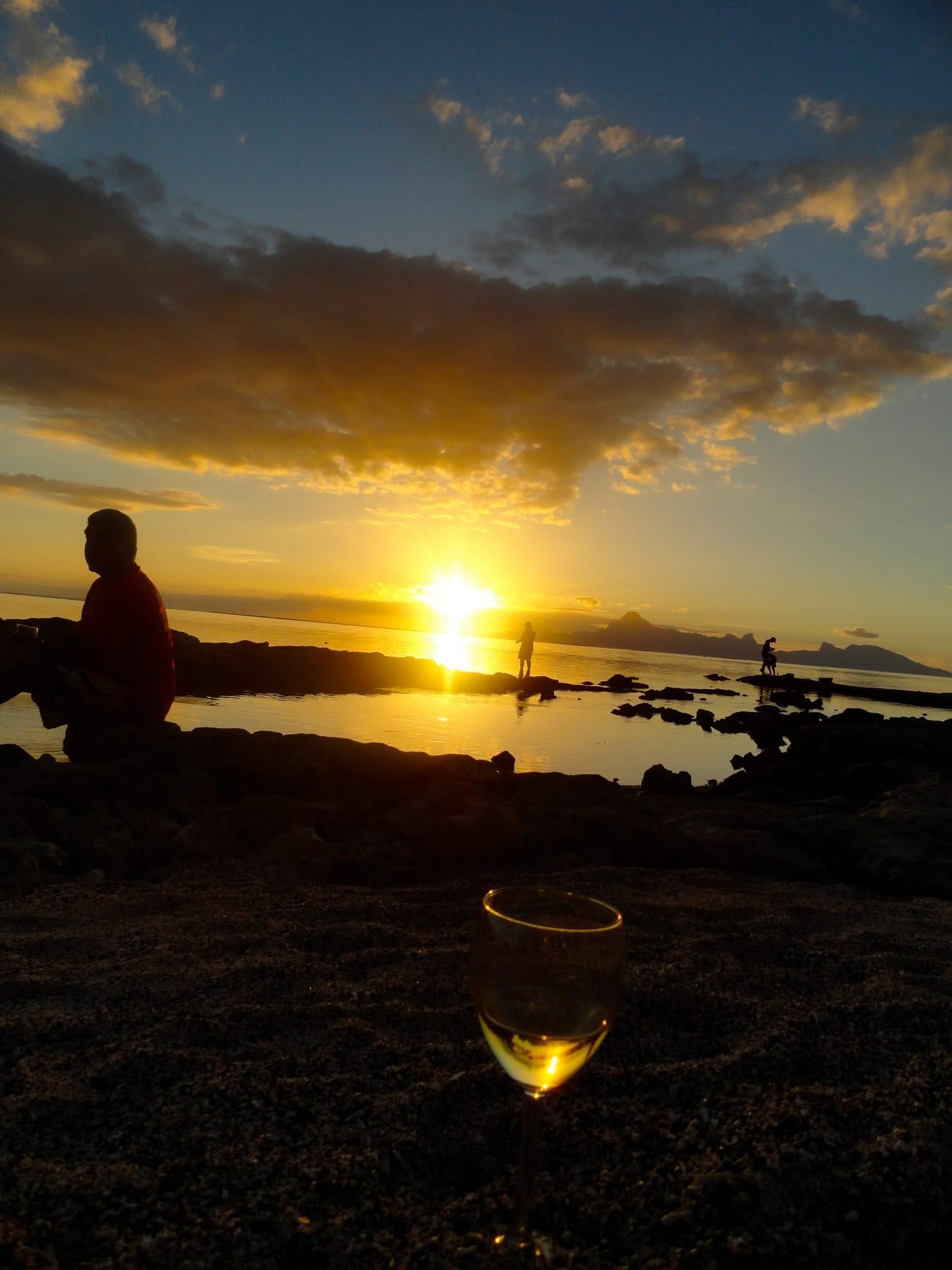 Un verre posé sur le sable noir, le soleil se couche derrière Moorea